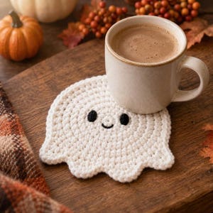 Puede incluir: Un posavasos de ganchillo blanco con forma de fantasma, con dos pequeños ojos negros bordados y una boca negra curvada, colocado bajo una taza de cerámica beige con una bebida marrón, con superficie texturizada y bordes redondeados.
