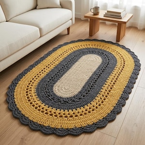 May include: An oval-shaped crochet rug featuring concentric bands of cream, dark gray, and mustard yellow yarn. The rug has a central solid cream section, a textured dark gray middle band, and a wide mustard yellow border with a scalloped edge.