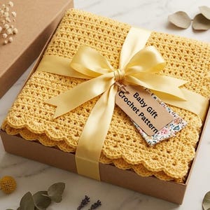 May include: A yellow crochet baby blanket with a scalloped edge, tied with a yellow satin ribbon, presented in a brown gift box. A tag that reads "Baby Gift Crochet Pattern" is attached to the ribbon. The blanket is approximately 30 inches square.
