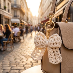 May include: A beige handbag with a gold-coloured keyring featuring a crocheted bow. The bow is cream-coloured and has an openwork design. The background shows a European street scene with people and buildings.