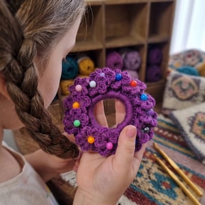May include: A purple crochet hair scrunchie adorned with small, crocheted flowers and colorful beads. The scrunchie is held in a person's hand, with a braided hairstyle visible in the background. Crochet hooks and yarn are also in the background.
