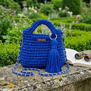 May include: A vibrant royal blue crochet handbag featuring a thick braided handle, a matching blue tassel attached to a silver ring, and a long chain strap woven with blue fabric. A small rectangular brown label is attached to the front centre.
