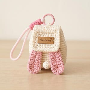 May include: A cream-colored crochet backpack keychain featuring two pink rabbit ears, a small white pom-pom tail, and a pink plastic ring attachment. A rectangular brown leather tag with the word Handmade and a small heart is centered on the flap.
