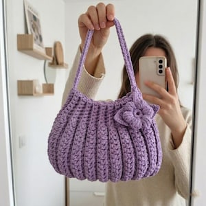 May include: A handmade, lavender crochet handbag with a flower accent. The bag features a ribbed design and two handles. The bag is held up in front of a mirror.