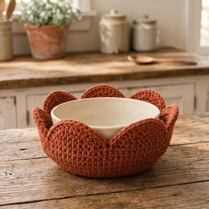 May include: A round, off-white ceramic bowl nestled inside a rust-coloured, crocheted fabric bowl cosy. The cosy features a scalloped, petal-like edge design that wraps around the exterior of the bowl, providing a textured, thick yarn layer.