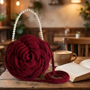 May include: A deep red crochet handbag shaped like a large blooming rose. The bag features a circular, layered petal design and a single handle made of white spherical faux pearls. A long, matching red crochet strap is attached to the side.