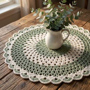 May include: A round, crocheted doily featuring alternating concentric rings of sage green and cream yarn. The doily has a scalloped edge and supports a small, off-white ceramic pitcher filled with green eucalyptus branches and leaves.