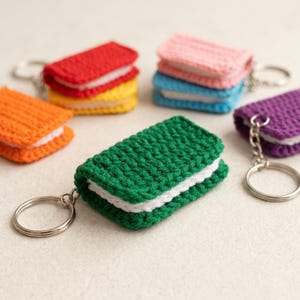 May include: A collection of rectangular crochet keychains in various colors including green, orange, red, yellow, pink, blue, and purple. Each keychain features a white center layer resembling a sandwich cookie and a silver metal keyring attached.