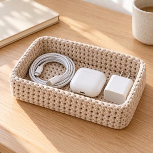 May include: A rectangular beige crochet basket containing a white coiled USB-C charging cable, a white wireless earbud charging case, and a white wall power adapter. The basket features a thick, textured knit pattern made of yarn.