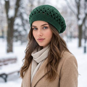 Puede incluir: Una boina de ganchillo verde oscuro con un patrón calado texturizado en la corona y una banda acanalada sólida. El gorro se lleva con una bufanda beige claro y un abrigo beige, ofreciendo una vista clara del intrincado trabajo de hilo.