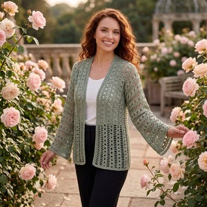 May include: A sage green open-front cardigan featuring an intricate crochet knit pattern with long, flared sleeves. The garment is worn over a simple white tank top and black trousers, showcasing a textured, lightweight design.