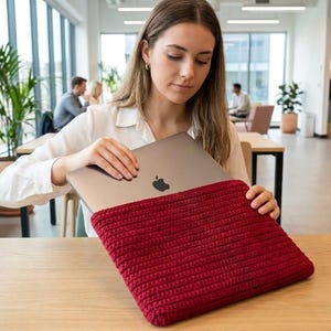 Puede incluir: Una funda para portátil rectangular de ganchillo en color rojo oscuro con un patrón de puntos acanalados y texturizados. La funda está diseñada para albergar un portátil gris metálico fino con un logotipo de Apple negro en el centro.