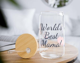 May include: A clear glass can-shaped tumbler with the text "World's Best Mama!" and floral accents. It includes a bamboo lid and a clear straw, resting on a wooden table. The design is suitable for beverages.