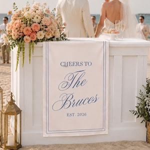 May include: A white rectangular wedding sign featuring a blue double-line border and blue text that reads CHEERS TO The Bruces EST. 2026. The sign is draped over the front of a white wooden bar counter decorated with a large floral arrangement.