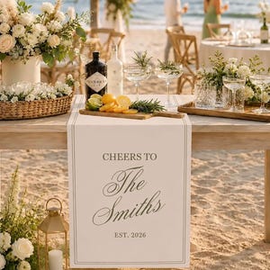 Peut inclure: Un chemin de table en tissu blanc portant l'inscription imprim&eacute;e "CHEERS TO The Smiths EST. 2026" dans une police serif et cursive gris fonc&eacute;. Le chemin est pos&eacute; sur une table en bois avec un arrangement floral, une bouteille de gin et des verres.