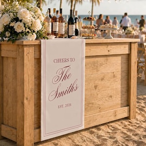 May include: A rectangular white fabric runner draped over a wooden bar, featuring the maroon text "CHEERS TO The Smiths EST. 2026" with a double-line border. The runner is positioned on a rustic wooden bar counter holding wine bottles and flowers.