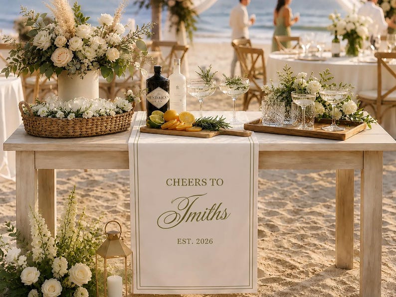 Peut inclure: Un chemin de table blanc avec le texte imprim&eacute; CHEERS TO Smiths EST. 2026, pos&eacute; sur une table en bois. La table est d&eacute;cor&eacute;e d'un arrangement floral dans un panier, d'une bouteille de gin, d'agrumes tranch&eacute;s, de romarin et de verres sur des plateaux.
