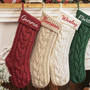 May include: Four cable-knit Christmas stockings hanging in a row. The first is deep red with the name Gemma, the second is beige with the name Benson, the third is cream with the name Wesley, and the fourth is dark green with the name Martin.