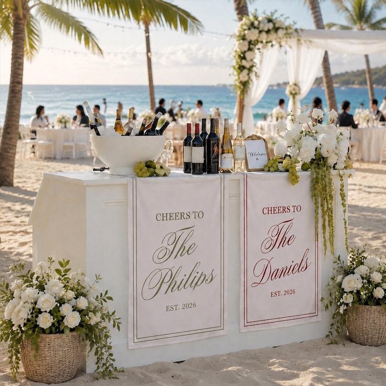 May include: A white bar counter on a sandy beach featuring two hanging banners. One banner reads Cheers to The Philips Est. 2026, and the other reads Cheers to The Daniels Est. 2026. The bar is decorated with white floral arrangements and wine bottles.