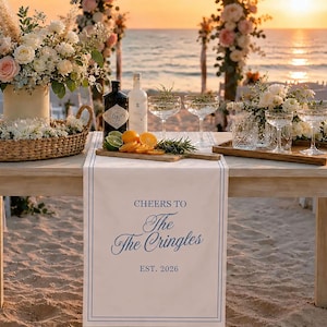 May include: A white table runner with blue border lines and the printed text CHEERS TO The The Cringles EST. 2026. The runner is displayed on a wooden table set with floral arrangements, a wicker basket, glass coupes, and a wooden cutting board.