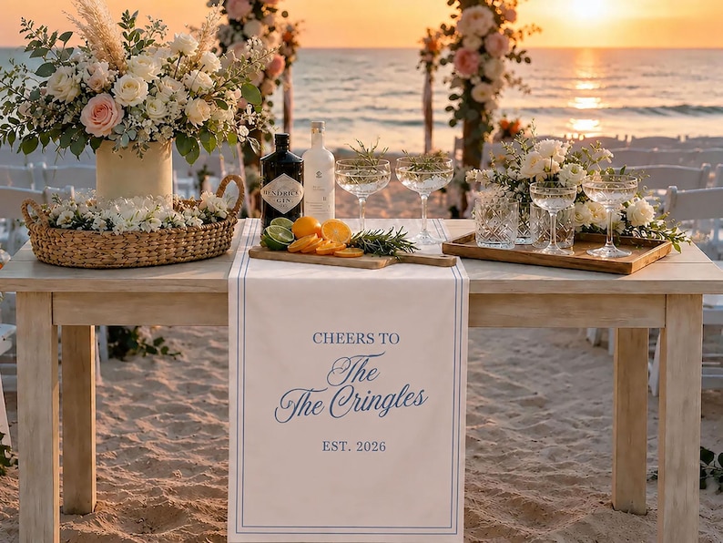 May include: A white table runner with blue border lines and the printed text CHEERS TO The The Cringles EST. 2026. The runner is displayed on a wooden table set with floral arrangements, a wicker basket, glass coupes, and a wooden cutting board.