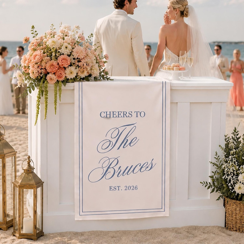 May include: A white rectangular wedding sign featuring a blue double-line border and blue text that reads CHEERS TO The Bruces EST. 2026. The sign is draped over the front of a white wooden bar counter decorated with a large floral arrangement.