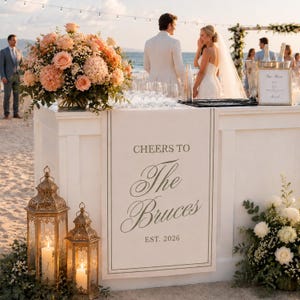 Peut inclure: Un panneau de bar de mariage blanc portant l'inscription 'CHEERS TO The Bruces EST. 2026' en gris foncé. Le panneau est présenté sur un comptoir blanc décoré d'une grande composition florale pêche, de deux lanternes dorées et de verres.