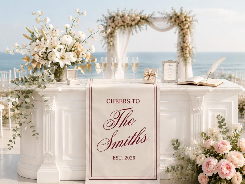 May include: A white wedding table runner featuring the maroon text CHEERS TO The Smiths EST. 2026, draped over a white altar table. The table is decorated with white floral arrangements, four champagne flutes, a ring box, and an open guest book.