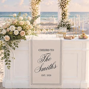 Peut inclure: Un bar de mariage blanc avec un chemin de table portant l'inscription CHEERS TO The Smiths EST. 2026. Le bar est orn&eacute; d'un grand arrangement de roses blanches et roses, quatre verres &agrave; vin, une bo&icirc;te en verre, un livre et des macarons.