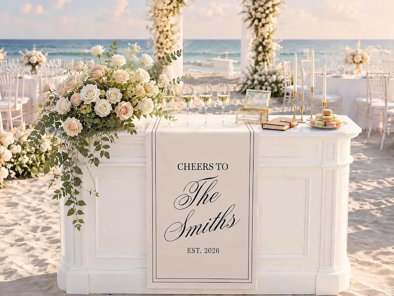 Peut inclure: Un bar de mariage blanc avec un chemin de table portant l'inscription CHEERS TO The Smiths EST. 2026. Le bar est orn&eacute; d'un grand arrangement de roses blanches et roses, quatre verres &agrave; vin, une bo&icirc;te en verre, un livre et des macarons.