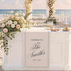 May include: A white wedding bar featuring a runner with the text CHEERS TO The Smiths EST. 2026. The bar is decorated with a large arrangement of white and blush roses, four clear wine glasses, a glass box, a small book, and a plate of macarons.