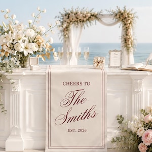 Peut inclure: Un chemin de table de mariage blanc avec le texte bordeaux CHEERS TO The Smiths EST. 2026, drap&eacute; sur une table d'autel blanche. La table est d&eacute;cor&eacute;e de compositions florales blanches, quatre fl&ucirc;tes &agrave; champagne, un &eacute;crin et un livre d'or ouvert.