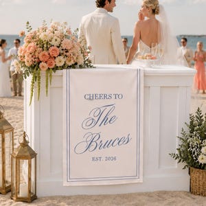 Peut inclure: Une pancarte de mariage rectangulaire blanche avec une bordure bleue &agrave; double ligne et un texte bleu indiquant CHEERS TO The Bruces EST. 2026. La pancarte est drap&eacute;e sur le devant d'un comptoir de bar en bois blanc orn&eacute; de fleurs.