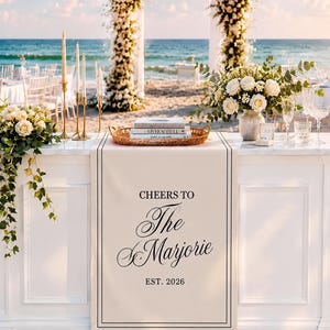 Peut inclure: Un chemin de table rectangulaire blanc avec le texte imprim&eacute; en noir CHEERS TO The Marjorie EST. 2026. Le chemin est pos&eacute; sur une table blanche sur une plage de sable, orn&eacute; de compositions florales, de bougies et de livres dans un plateau.