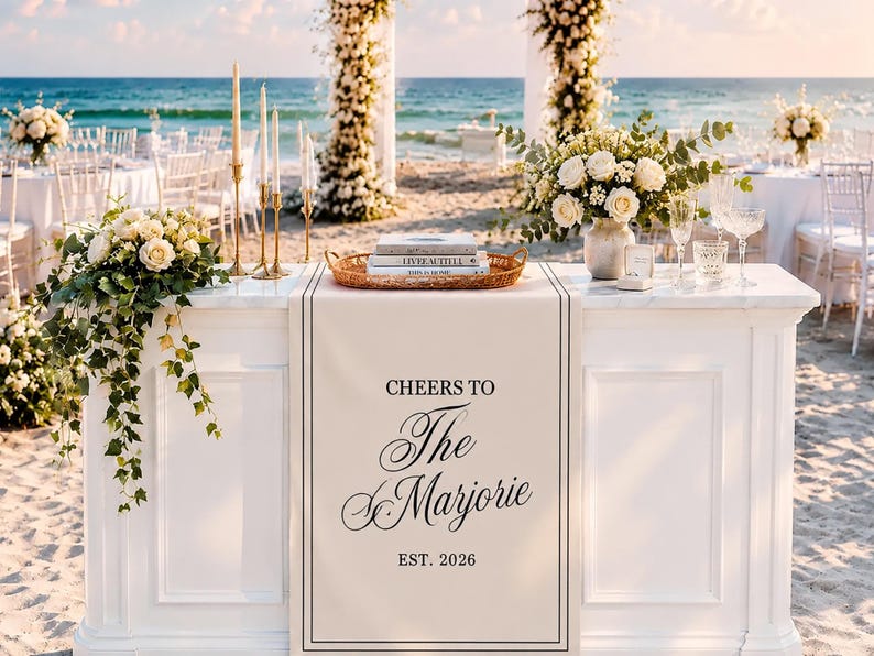 Peut inclure: Un chemin de table rectangulaire blanc avec le texte imprim&eacute; en noir CHEERS TO The Marjorie EST. 2026. Le chemin est pos&eacute; sur une table blanche sur une plage de sable, orn&eacute; de compositions florales, de bougies et de livres dans un plateau.
