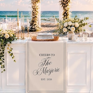 May include: A white rectangular wedding table runner featuring the black printed text 'CHEERS TO The Marjorie EST. 2026' centered within a thin black border. The runner is draped over a white table decorated with floral arrangements, candles, and books.