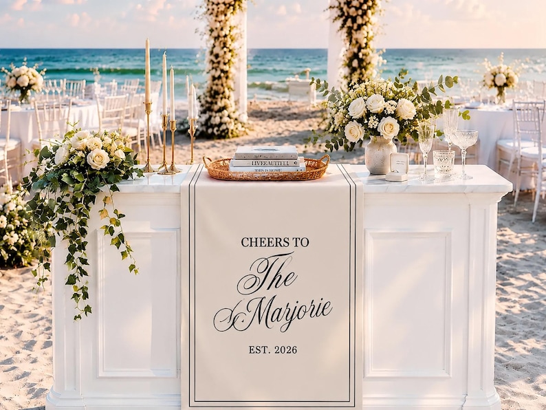 May include: A white rectangular wedding table runner featuring the black printed text 'CHEERS TO The Marjorie EST. 2026' centered within a thin black border. The runner is draped over a white table decorated with floral arrangements, candles, and books.