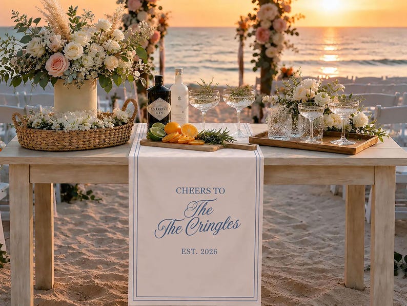 Peut inclure: Un chemin de table blanc avec des bordures bleues et le texte imprim&eacute; CHEERS TO The The Cringles EST. 2026. Le chemin est pos&eacute; sur une table en bois avec des compositions florales, un panier en osier, des coupes en verre et une planche.