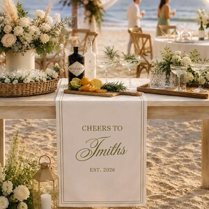 May include: A white fabric table runner featuring the text "CHEERS TO Smiths EST. 2026" in dark lettering, draped over a wooden table. The table is decorated with floral arrangements, a bottle of gin, citrus slices, and glassware for a celebration.