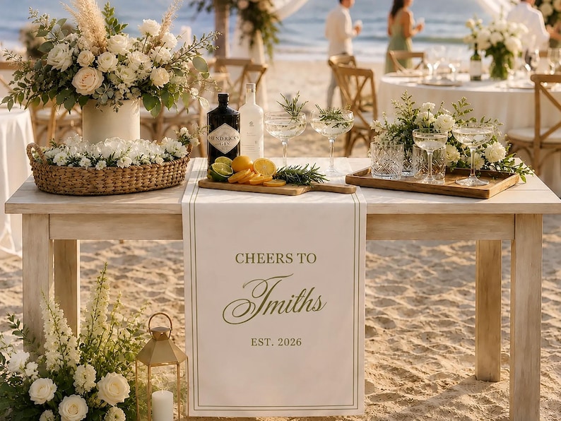 May include: A white fabric table runner featuring the text "CHEERS TO Smiths EST. 2026" in dark lettering, draped over a wooden table. The table is decorated with floral arrangements, a bottle of gin, citrus slices, and glassware for a celebration.
