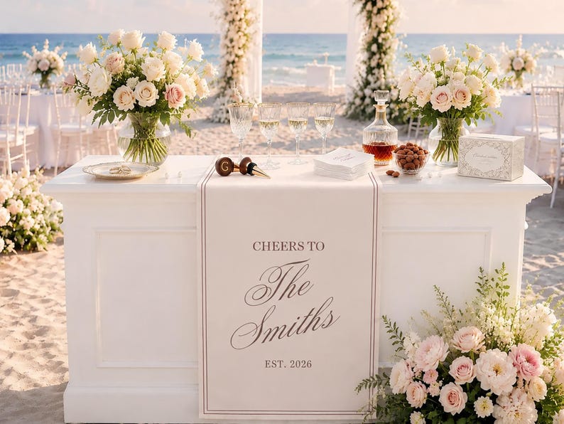 Peut inclure: Une table de bar de mariage blanche avec un chemin de table personnalis&eacute; portant le texte CHEERS TO The Smiths EST. 2026. La table est orn&eacute;e de deux grands bouquets de roses blanches et roses, une carafe en verre, quatre verres et des serviettes.