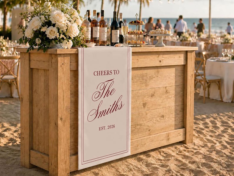 Peut inclure: Un bar rectangulaire en bois avec un chemin de table blanc drap&eacute; sur le devant. Le tissu affiche le texte bordeaux "CHEERS TO The Smiths EST. 2026" encadr&eacute; par de fines lignes verticales. Le bar est surmont&eacute; de bouteilles de vin et de fleurs.