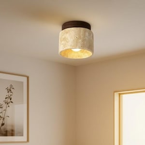 May include: A bedroom scene featuring a low wooden bed frame with white bedding, a framed botanical print, and a small plant. A textured, cylindrical ceiling light with a brown top and gold interior illuminates the room. The room has a minimalist design.