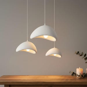 Peut inclure: Trois lampes suspendues blanches et texturées, de forme incurvée et organique, sont suspendues au plafond. Chaque lampe est équipée d'une ampoule chaude et brillante. Les lampes sont suspendues au-dessus d'une table en bois.