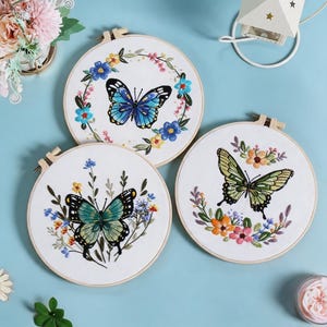 May include: Three round embroidery hoops with butterfly designs. Each hoop features a white fabric background with a butterfly in the center, surrounded by colorful floral embroidery. The butterflies are in shades of blue, green, and yellow.