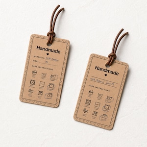 Puede incluir: Dos etiquetas rectangulares de papel color canela con esquinas redondeadas y cordones marrones. Cada etiqueta muestra la palabra Handmade, un corazón, campos de material y talla, y una cuadrícula de iconos de cuidado en negro.