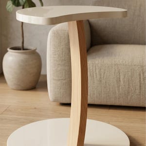 May include: A side table with a glossy off-white kidney-shaped top and base, connected by a single curved light-toned wooden support pillar. The base features the text NEXORA with a blue and white abstract logo in the bottom left corner.
