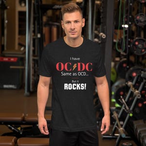 Può includere: T-shirt grigio scuro con la scritta "I have OC⚡DC Same as OCD... But it ROCKS!" in rosso e bianco. La maglietta è a maniche corte e ha lo scollo tondo. La persona è in palestra.