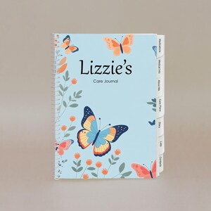 May include: A spiral-bound care journal with a light blue cover featuring the name "Lizzie's" and the words "Care Journal". The cover is decorated with colorful butterflies and floral elements. The journal has labeled divider tabs.