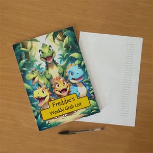 Puede incluir: Un bloc de notas semanal con una colorida ilustración de cuatro dinosaurios de dibujos animados en una selva. Una etiqueta rectangular amarilla en la parte frontal muestra el texto Freddie's Weekly Grab List. Un bolígrafo negro yace debajo.
